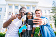 © Iryna - Happy interracial boys and african girl friend posing for cute selfie.Young friends in t-shirts photographing together against university building. Relationship multhetnic friendship concept.