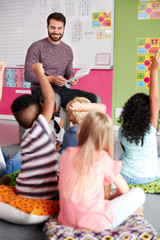 Wall Mural - Elementary Pupils Raising Hands To Answer Question As Male Teacher Reads Story In Classroom