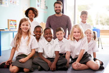 Wall Mural - Portrait Of Elementary School Pupils Wearing Uniform Sitting On Floor In Classroom With Male Teacher