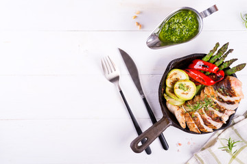 Wall Mural - Chicken breast grill with bbq vegetables and pesto sauce in a cast iron pan on a white background, flat lay,