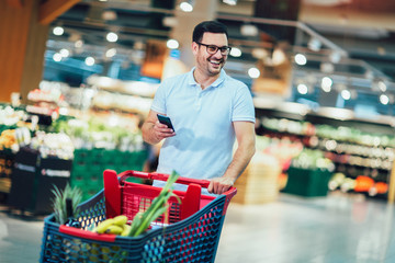 Handsome man shopping in supermarket pushing trolley and holding phone.