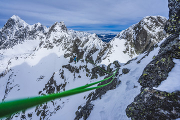 Wall Mural - An alpinist climbing on a winter rocky ridge in high alpine like winter mountain landscape. Climber or mountainer on a ridge, green climbing rope flying in the wind.