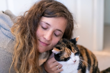 Closeup portrait of happy smiling young woman bonding with calico cat pet companion, bumping rubbing bunting heads, friends showing affection