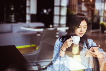 Asian woman with beautiful smile hold mobile phone during rest in coffee shop.Attractive woman drinking coffee.Reflection glass window.