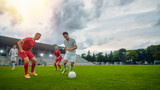 Fototapeta Sport - Professional Soccer Player Leads with a Ball, Masterfully Dribbling and Bypassing Sliding Tackles of His Opponents. Two Professional Football Teams Playing. Low Angle Shot. Warm Sunlight Flare.