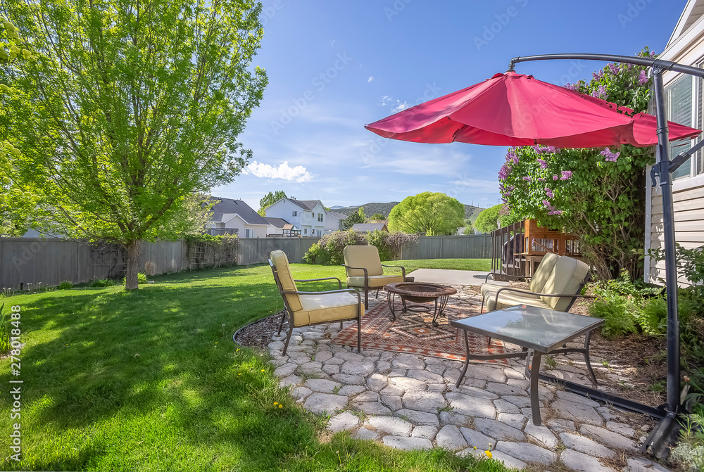 Seating area on a stone patio under the shade of a red umbrella on a sunny day - obrazy, fototapety, plakaty 