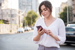 © AnnaDemy - Beautiful brunette business woman in white skirt and grey suit trousers working on a mobile phone in her hands outdoors. European city on background. copy space