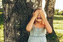 Horizontal Outdoor Image Of Smiling Little Girl Covering Her Eyes With Both Hands, Playing Hide And Seek Standing Next A Big Tree. Adorable Child Having Fun Outdoors In The Park. Childhood