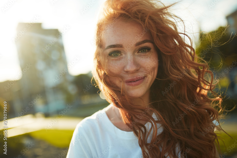 Portraits of a charming red-haired girl with a cute face. Girl posing for the camera in the city center. She has a wonderful mood and a lovely smile - obrazy, fototapety, plakaty 