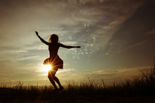 Silhouette Of A Girl In A Dress On The Background Of The Sunset Sky And The Sun, Below - The Grass. She Is Dancing. The Concept Of Freedom, Happiness.