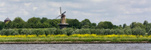 View On The Shore Of The Merwede River In The Netherlands, Typical Dutch Landscape With Traditional Windmill To Provide Energy And Power. Real Dutch Weather With Wind, Clouds And Sun.