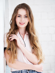 Lifestyle portrait beautiful blonde woman in pink blouse long wavy hair leaning on white wall looking into camera with adorable smile, natural light slide film colors