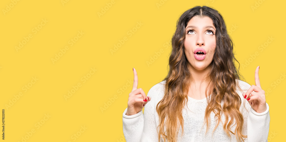 Young beautiful woman wearing white sweater amazed and surprised looking up and pointing with fingers and raised arms.