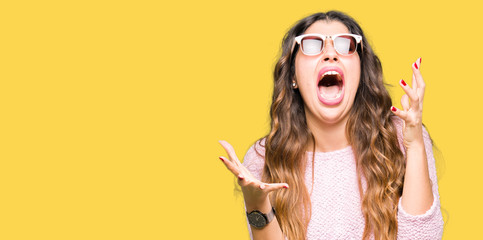 Sticker - Young beautiful woman wearing sunglasses and pink sweater crazy and mad shouting and yelling with aggressive expression and arms raised. Frustration concept.