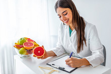 Portrait Of Young Smiling Female Nutritionist In The Consultation Room. Nutritionist Desk With Healthy Fruit, Juice And Measuring Tape. Dietitian Working On Diet Plan.