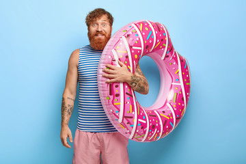 Wall Mural - Portrait of carefree smiling red haired man wears sailor vest, pink shorts, carries rubber swimring, going to swimming pool, has good mood, isolated on blue background. Recreation time concept