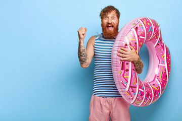 Yeah, finally summer! Joyful foxy holidaymaker clenches fist, rejoices coming holiday, dressed in casual outfit, carries inflated donut ring, shouts from pleasure, wins ticket for tour abroad