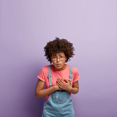 Wall Mural - Upset depressed woman with Afro hairstyle, keeps hands near chest, suffers from heart attack, has problems with health, has embarrassed expression, needs doctors help, wears t shirt and denim sarafan