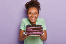 Overjoyed Curly Dark Skinned Girl Holds Birthday Cake, Smiles Broadly, Bakes Tasty Dessert On Special Occasion, Dressed Casually, Grins At Camera, Shows White Teeth, Models Over Purple Studio Wall