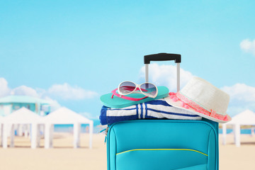 holidays. travel concept. blue suitcase with female hat, flip flips, sunglasses and beach towel in front of tropical background