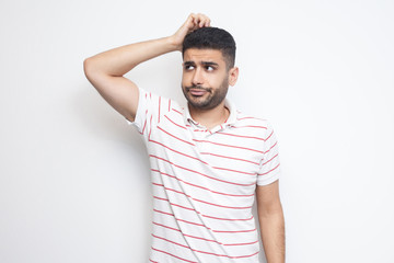 Wall Mural - Portrait of query handsome bearded young man in striped t-shirt standing scratching his head, planing and thinking about something. indoor studio shot, isolated on white background.