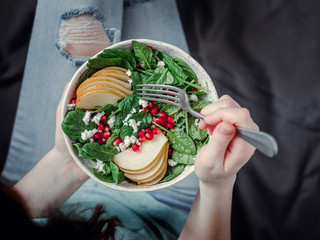 Woman in jeans holding vegan salad bowl with spinach, pear, pomegranate, cheese. Vegan breakfast, vegetarian food, diet concept. Girl in jeans holding fork with knees and hands visible