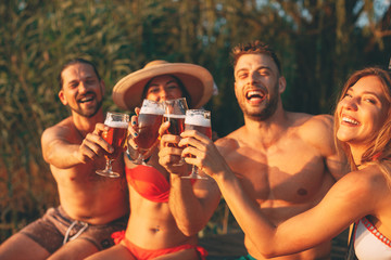 Sticker - Happy group of young people toasting with beer on a dock by the river during the summer sunny day. Selective focus on beer