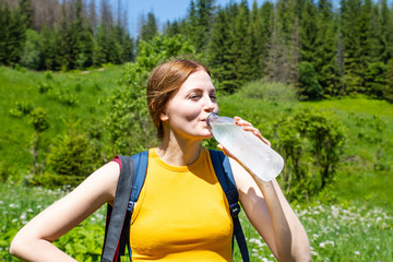 Female tourist girl in a yellow T-shirt and blue jeans shorts drink water from a plastic bottle on a background of green forest .Woman resting in the mountains. Travel and tourism concept