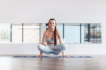 Canvas Print - Positive young woman yoga lover doing kukkutasana sitting in white gym. Concept of regular exercise, healthy lifestyle. Arm balance exercise with crossed legs, Scale Posture, Tolasana, Utpluthi Pose