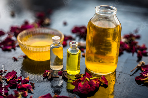 Close up of castor oil, tea tree oil, and some coconut oil in bottles on the wooden surface along with some raw honey and rose petals also present on the surface.