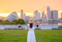 Young Woman Traveling With Hat At Sunset, Happy Asian Traveler Visit In Singapore City Downtown. Landmark And Popular For Tourist Attractions. Asia Travel Concept