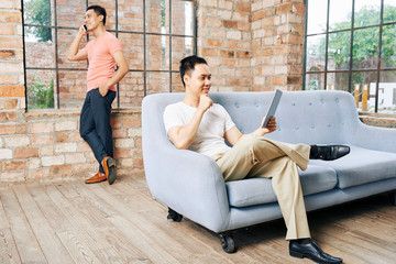 Wall Mural - young vietnamese man resting on sofa and reading article on tablet computer when his coworker talkin