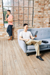 Wall Mural - positive entrepreneur sitting on sofa and watching webinar on tablet computer when his coworker maki