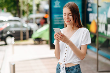 Smiling  young woman holding smartphone and looking at the screen standing outdoor- Image