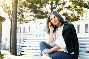 Style, age, people and modern electronic devices concept. Trendy looking happy female in her fifties relaxing on bench after long walk, sitting in comfortable relaxed pose, talking on mobile phone