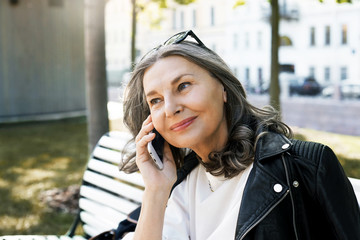 Wall Mural - Always in touch. Outdoor view of attractive fashionable middle aged woman in stylish leather jacket having nice phone conversation in park. Positive joyful mature female speaking on mobile