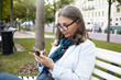 © shurkin_son - Leisure, technology, business and modern lifestyle concept. Serious gray haired mature woman in stylish rectangular eyewear sitting on bench in park, using smart phone, messaging via online app