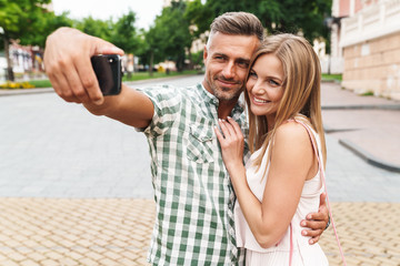 Wall Mural - Image of pleased young couple smiling and taking selfie photo together while walking through city street