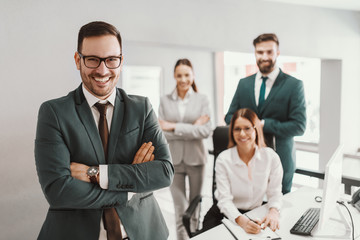 Young handsome smiling CEO in suit standing in office with arms crossed. In background his successful team posing.