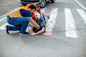 Wall Mural - Medic with car driver applying first aid to the injured man lying on the pedestrian crossing after the road accident