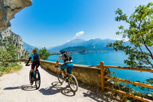 Cycling Woman And Man Riding On Bikes At Sunrise Mountains And Garda Lake Landscape. Couple Cycling MTB Enduro Flow Sentiero Ponale Trail Track. Outdoor Sport Activity.