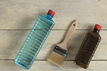 A brush lies next to plastic bottles with stain and solvent on an old white vintage wooden plank table. Place for text or logo.