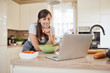© nenadaksic - Pretty Caucasian brunette in apron leaning on kitchen counter and looking at laptop and following recipe for dinner. Woman holding carrot in hand.