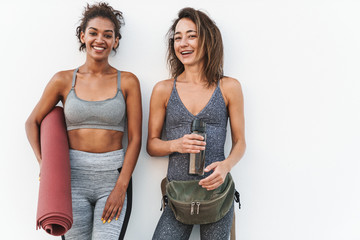 Poster - Photo of two young multiethnic sportswomen smiling and holding yoga mat while standing over white wall outdoors