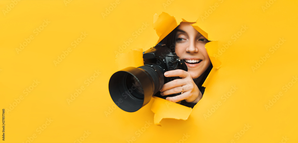 A young paparazzi girl holds a reflex camera and looks through a torn hole in yellow paper. The concept of smile and laughter from what he saw. Copy space. - obrazy, fototapety, plakaty 