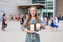 Young Teen Girl/Middle School Student Standing In Front Of School With Awards And Diploma After Grade 8/Middle School Graduation Ceremony With Blurred People In Background.