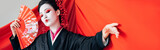beautiful geisha in black kimono with hand fan and red cloth on background gesturing isolated on white, panoramic shot