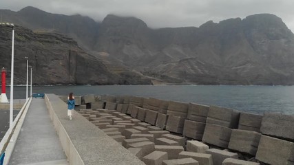 Wall Mural - Aerial view. Hipster girl walks along a long concrete pier on the background of large and beautiful mountains covered with clouds. Agaete, Gran Canaria