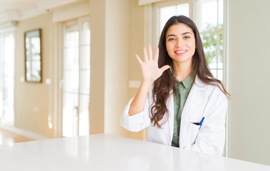 Canvas Print - Young woman wearing medical coat at the clinic as therapist or doctor showing and pointing up with fingers number five while smiling confident and happy.