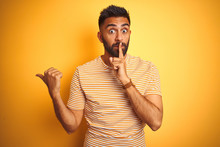 Young Indian Man Wearing T-shirt Standing Over Isolated Yellow Background Asking To Be Quiet With Finger On Lips Pointing With Hand To The Side. Silence And Secret Concept.